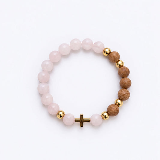 A beaded stretch bracelet featuring light pink rose quartz beads, natural brown wood beads, polished gold spacer beads, and a small gold cross charm, displayed on a clean white background.