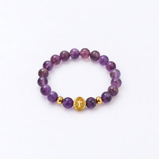 A purple gemstone beaded bracelet with two small gold spacer beads and a central gold round charm featuring a St Benedict symbol, displayed on a clean white background.