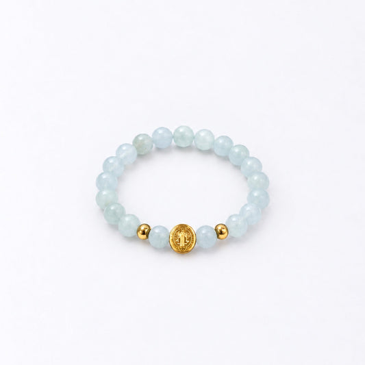 A beaded stretch bracelet featuring light blue-green round beads, two small gold spacer beads, and a central gold round charm with a St Benedict symbol, displayed on a clean white background.